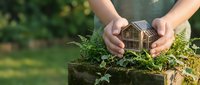 Hands protecting wooden house model on moss-covered surface with green plants, symbolizing home insurance, real estate protection and environmental care.