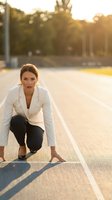 Professional businesswoman in white blazer crouched at starting line on running track during golden hour sunset, ready to sprint forward in athletic stance.