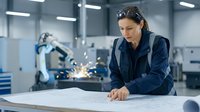 Female engineer of South Asian descent in dark blue work jacket examines blueprints at industrial workstation with robotic arm in background, representing manufacturing and automation technology.