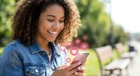 Young African American woman with curly hair smiling while using smartphone outdoors, receiving social media likes and notifications in urban park setting.