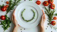 Female hands holding white plate with rosemary wreath, surrounded by fresh cherry tomatoes and herbs on light background. Food styling composition.