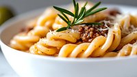 Creamy pasta dish with ground meat sauce garnished with fresh rosemary sprig in white ceramic bowl, close-up view showing texture of spiral noodles and meat.