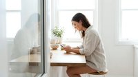 Young woman with dark hair and light skin tone, wearing casual linen shirt, writing or taking notes at white desk by bright window with potted plant, natural daylight interior workspace.