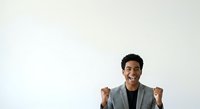 Young male professional with dark hair and medium skin tone wearing a gray blazer, smiling with fists raised in celebration gesture against white background for success and achievement concepts.