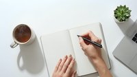 Hands writing in open notebook on white desk with coffee cup and succulent plant, top view workspace for planning and creativity.