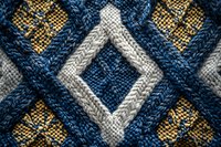 Close-up of knitted fabric with geometric diamond pattern in navy blue, white and gold colors showing detailed wool texture and traditional design.