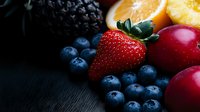 Fresh organic berries and fruits arranged on dark wooden background with dramatic lighting, featuring strawberries, blueberries, blackberries and citrus slices.