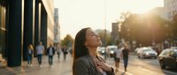 Young Caucasian businesswoman standing on busy city street at sunset, eyes closed, hands on chest, feeling calm and grateful.
