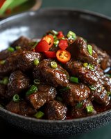Glazed Asian-style beef chunks garnished with fresh chili peppers and green onions in dark ceramic bowl, rich brown sauce with glossy finish for food service.