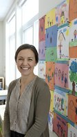 Happy elementary school teacher smiling in classroom with colorful student artwork display on wall behind her in bright educational environment.