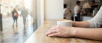 Woman's hands holding white coffee cup at wooden table in busy cafe with blurred customers and warm natural lighting creating cozy atmosphere for relaxation.