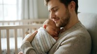 Young Caucasian father gently holds sleeping newborn baby against his chest, smiling softly in a cozy nursery room.