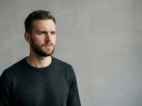 Young adult male with dark hair and beard wearing charcoal gray sweater against neutral gray background, suitable for corporate portraits, business profiles, and professional headshot applications.