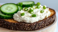 Fresh cottage cheese spread on dark rye bread garnished with green onions and cucumber slices, served on white ceramic plate. Healthy breakfast concept.