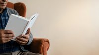Man with reddish-brown hair wearing a blue plaid shirt reads an open book while seated in a brown armchair against a neutral beige background.