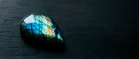 Polished labradorite gemstone with brilliant blue and green flash on dark slate surface for jewelry design and spiritual healing practices.