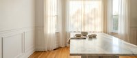 Bright modern kitchen interior with white marble countertop and wooden floors. Natural sunlight streaming through large windows creates warm atmosphere.