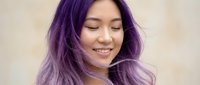 Young Asian woman with vibrant purple hair smiling with eyes closed, hair flowing in motion against neutral background for beauty and lifestyle concepts.