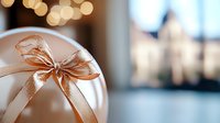 Elegant white Christmas ball ornament with shimmering rose gold bow against blurred festive bokeh background, creating soft dreamy holiday atmosphere.