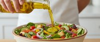 Pouring golden olive oil over fresh mixed green salad with tomatoes, cucumbers, and vegetables for cooking and food preparation.