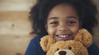 Cheerful African American child with curly hair hugging brown teddy bear, showing bright genuine smile against wooden background. Natural lighting creates warm atmosphere.