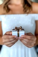 Female hands holding small white gift box with golden glitter bow against white dress. Romantic celebration and surprise concept for special occasions.