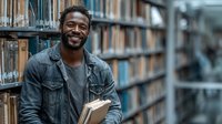 Smiling African American male student holding books in university library surrounded by tall bookshelves for education and academic research concepts.