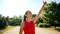 Young Asian woman in red athletic wear celebrating victory with raised fist outdoors on sunny day in park setting for fitness and wellness campaigns.