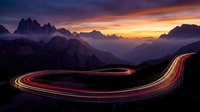 Mountain highway with light trails at sunset creating dramatic curves through alpine landscape with purple and orange sky.