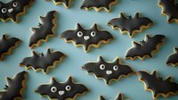 Dark chocolate bat-shaped cookies with white icing eyes arranged in rows on blue background, festive Halloween treats for party desserts and seasonal baking.