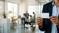 Businessman in dark blazer and light blue shirt holding blank white business card in modern office environment with colleagues and plants in background.