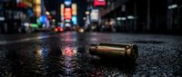 Spent brass bullet casing lies on wet urban street at night with blurred city lights and neon signs in background, depicting crime scene or danger.