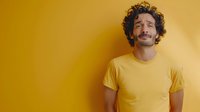 Young Hispanic man with mustache and curly hair wearing yellow t-shirt expressing peaceful emotion against yellow wall background, eyes closed and gentle smile.