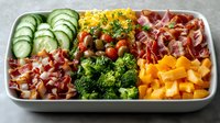 Charcuterie board with sliced cucumbers, cured meats, corn, broccoli, and bell peppers arranged in white rectangular tray for entertaining and catering.