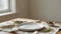 Minimalist table setting with empty white plate on beige linen placemat, fresh herbs and natural elements arranged for food styling and culinary design projects.