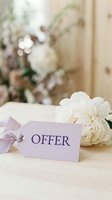 Elegant lavender gift tag with word Offer beside white peony flowers on wooden surface, soft bokeh floral background for wedding or retail promotion.