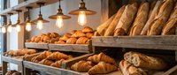 Rustic bakery display with fresh croissants, baguettes and pastries on wooden shelves under warm pendant lighting in artisan shop.
