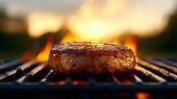 Juicy beef burger patty grilling on barbecue grate with flames and sunset background. Close up view shows seared meat texture and caramelization.