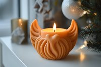 Orange decorative candle with wing design burning on white surface near Christmas tree with warm holiday lighting creating cozy festive atmosphere.