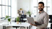 Young man with dark hair and beard, South Asian ethnicity, wearing casual button-up shirt, holding laptop while standing at modern office desk with plants and windows, smiling at camera in bright workspace.