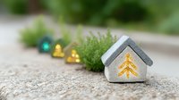 Small decorative winter house with golden tree pattern and colorful glass ornaments on stone surface with green plant, creating cozy holiday atmosphere.