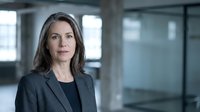 Middle-aged woman with long dark hair wearing dark blazer in modern office environment with glass partitions and natural light for corporate business portraits.