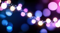 Glowing string lights create magical bokeh effect with pink, blue and warm white blurred circles against dark background, festive holiday atmosphere.