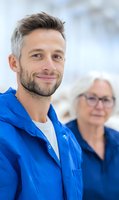 Caucasian male caregiver in blue uniform smiling warmly with senior patient in background, representing professional healthcare and elderly care services.