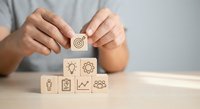 Business strategy concept with hands placing wooden blocks featuring target, lightbulb, gear, person, checklist, growth chart and team icons on desk.
