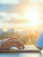 Female hands typing on laptop keyboard at sunset, urban skyline view through window, warm golden light creating atmospheric business workspace scene.