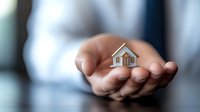 Small model house held gently in cupped hands against blurred background, representing real estate, property ownership, home protection, and housing market concepts.
