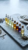 Assorted essential oils and aromatherapy serums in glass bottles with dropper caps arranged on slate surface with dried herbs and linen fabric for wellness and spa product marketing.