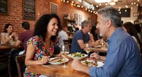 Happy mature couple enjoying romantic dinner date at trendy restaurant with brick walls and string lights creating warm intimate dining atmosphere.