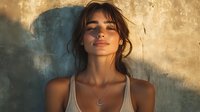 Young Hispanic woman with messy bun hairstyle wearing white tank top and necklace against textured wall in warm sunset lighting, eyes closed with serene expression.